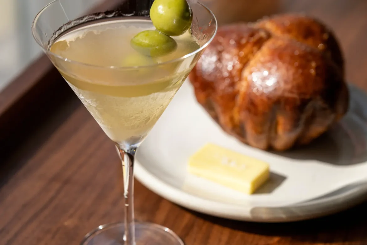 a brioche martini with olives on a dark wooden bar in front of a white plate with a brioche roll on it