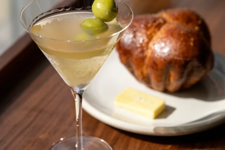 a brioche martini with olives on a dark wooden bar in front of a white plate with a brioche roll on it