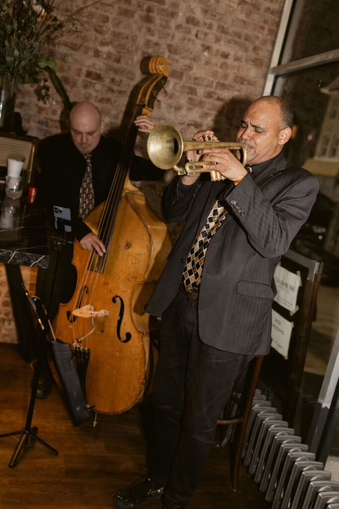 A trumpet player in front of a standing bass player in a room with a wood floor and exposed brick wall at the back